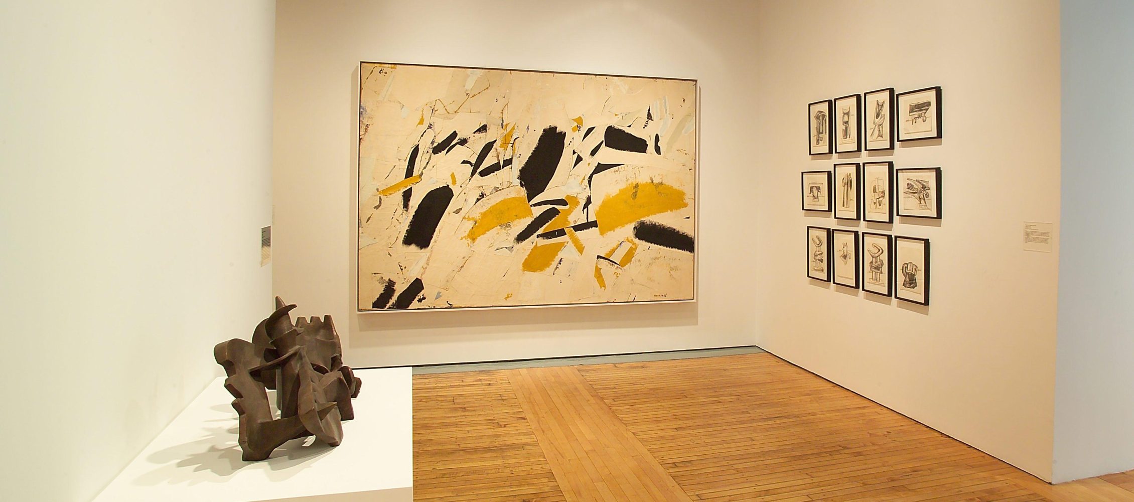 An art gallery interior with varied artworks on display, including framed paintings and drawings and a small sculpture on a pedestal.