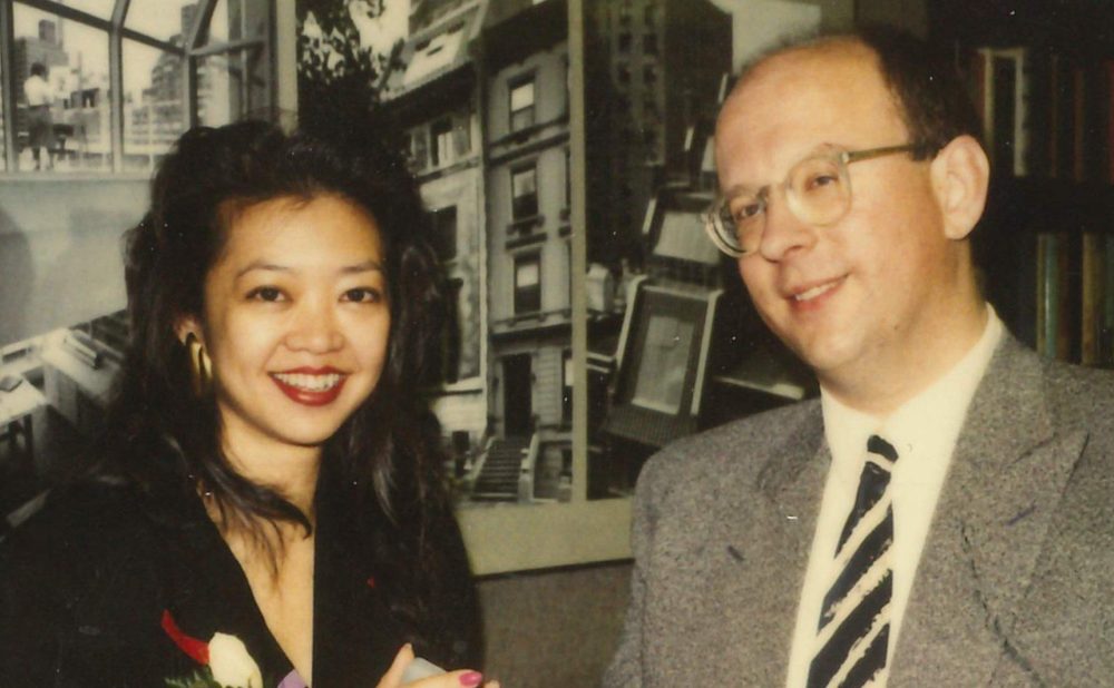 An Asian woman with long black hair a corsage and a white balding man with glasses in a gray suit standing together indoors.