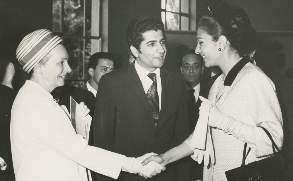 Black-and-white photograph of two women shaking hands, one in a headscarf, the other in gloves, with a man in a suit between them, indoors.
