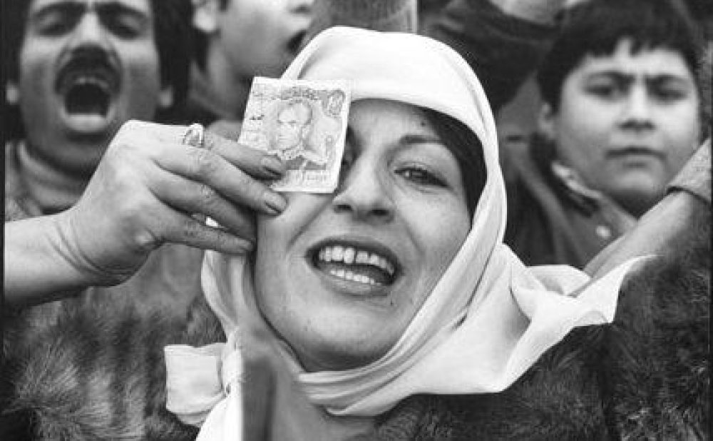 A black-and-white photo of a smiling woman in a hijab holding a banknote over one eye, surrounded by people.