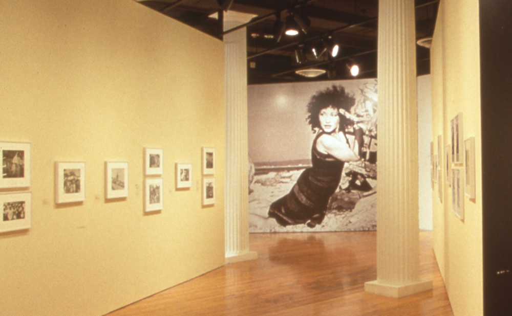 An art gallery with yellow walls, displaying framed photographs. A large black and white photo of a woman on a beach with windblown curly black hair is visible in the background. Alt-text generated by AI.