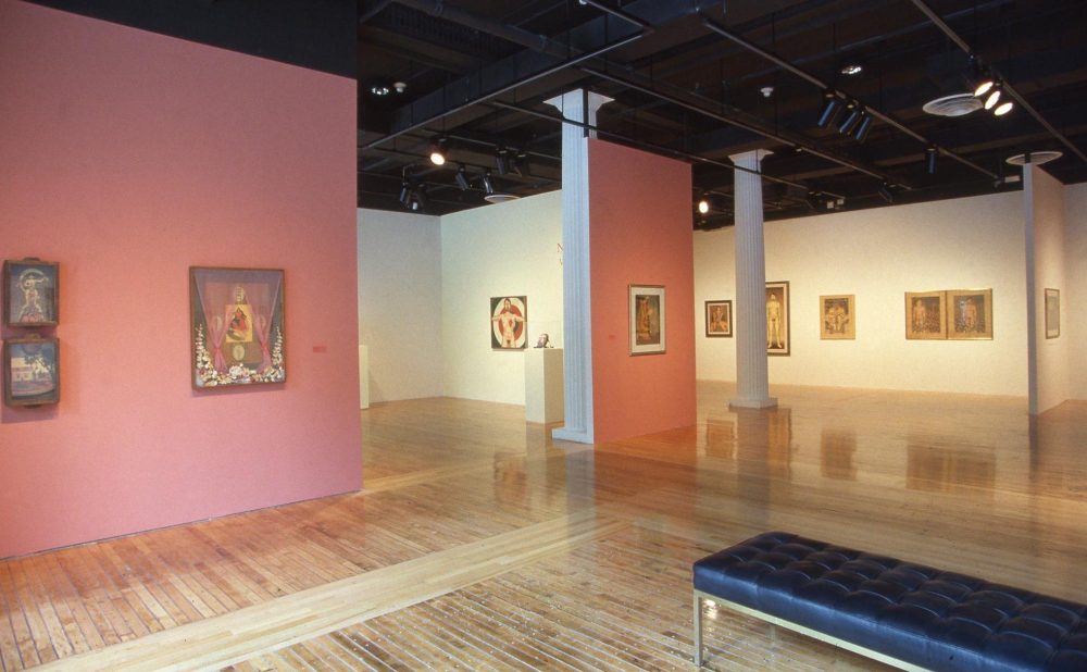 An art gallery with wooden floors, colorful walls, framed artworks, and a dark bench. Alt-text generated by AI.