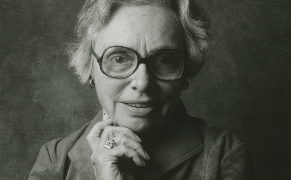 Black and white portrait of an elderly woman wearing glasses, her right hand resting on her chin. Alt-text generated by AI.