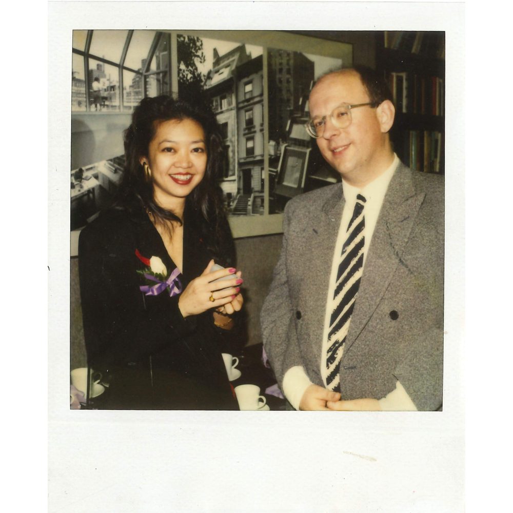 An Asian woman with long black hair a corsage and a white balding man with glasses in a gray suit standing together indoors.