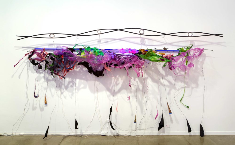 A sculpture comprising of a steel rod twisted in the shape of a DNA strand, hanging above a jubilant array of melted green, pink, and purple plastics and florescent lights. Tendrils of plastics reach down to the gallery floor.