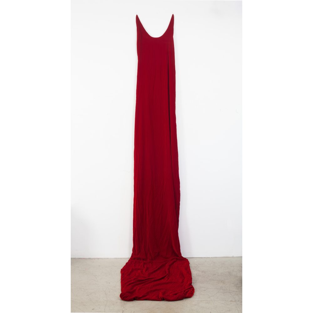 A sculpture of deep red velvet fabric takes the form of an absurdly long sleeveless dress; the shoulders of the dress are high up on the wall, and the body of the form cascades down the wall and pools onto the gallery floor.