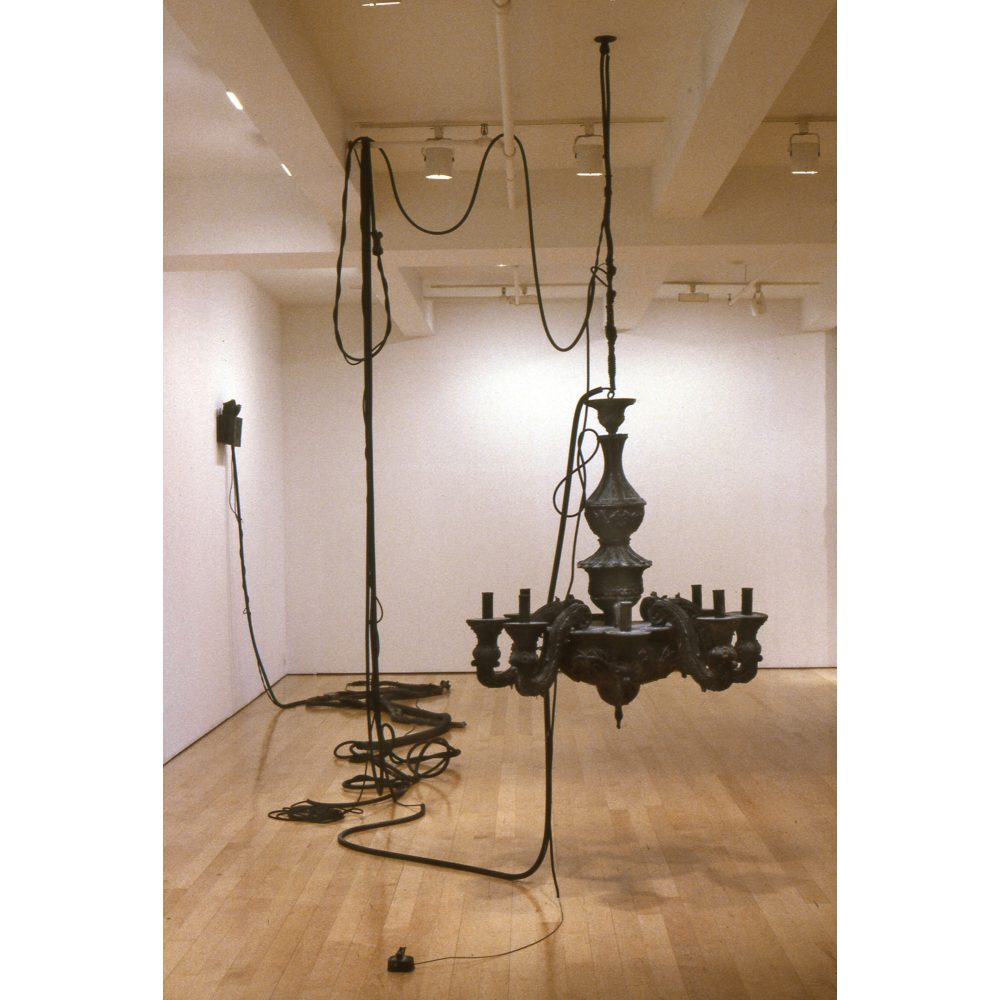 A black rubber installation fills a gallery, with a large suspended chandelier form as the central component, surrounded by carefully suspended and looped cords and faux electrical components, including an EXIT sign