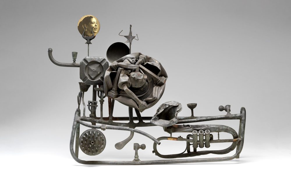 Sculptural assembly of metallic objects including a rose, pipes, and a portrait disk on a gray background.