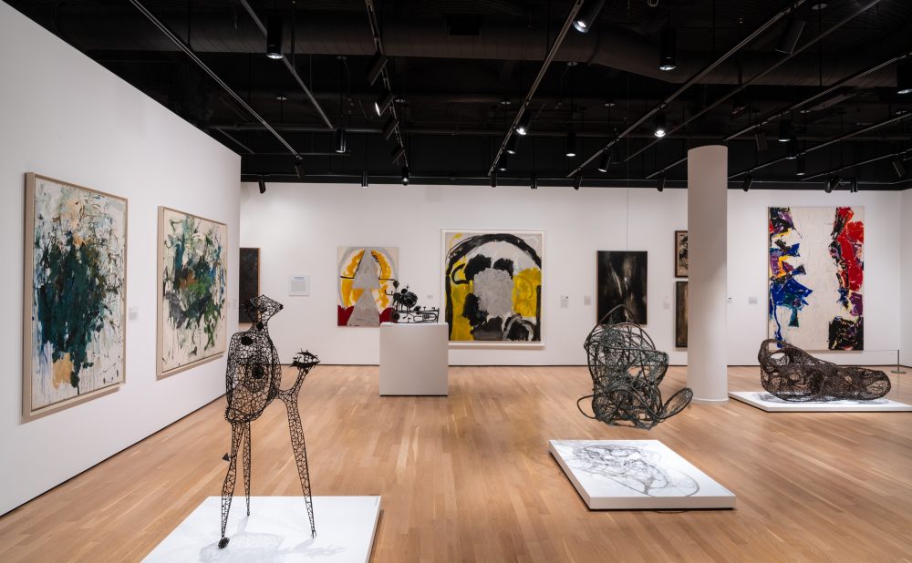 An art gallery featuring abstract paintings and sculptures on a wooden floor with bright lighting.