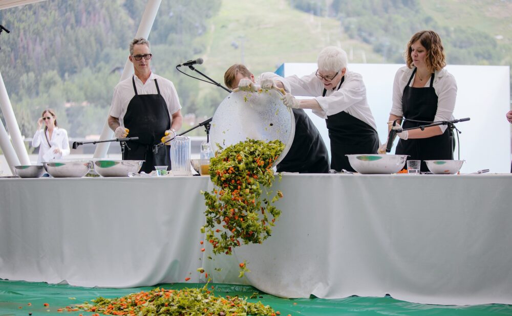 People in black aprons pour a large bowl of chopped salad over a table onto a green tarp during a cooking demonstration. Alt text generated by AI.