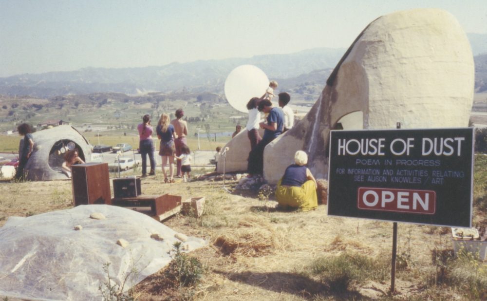 Outdoor art installation with people interacting near dome-like structures and a sign reading "House of Dust" against a hilly landscape. Alt-text generated by AI.  Transcribed Text:  HOUSE OF DUST POEM IN PROGRESS FOR INFORMATION AND ACTIVITIES RELATING SEE ALISON KNOWLES / ART OPEN