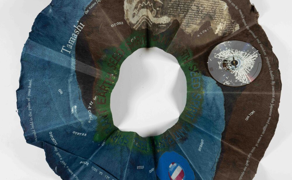 A circular, textured paper sculptural object, with gradients of blue and brown, featuring scattered text. Alt-text generated by AI.