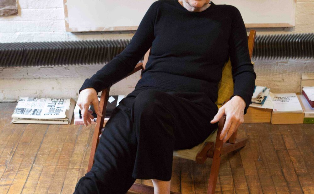 A person with white hair in black clothing seated on a wooden chair in an art studio. Alt-text generated by AI.