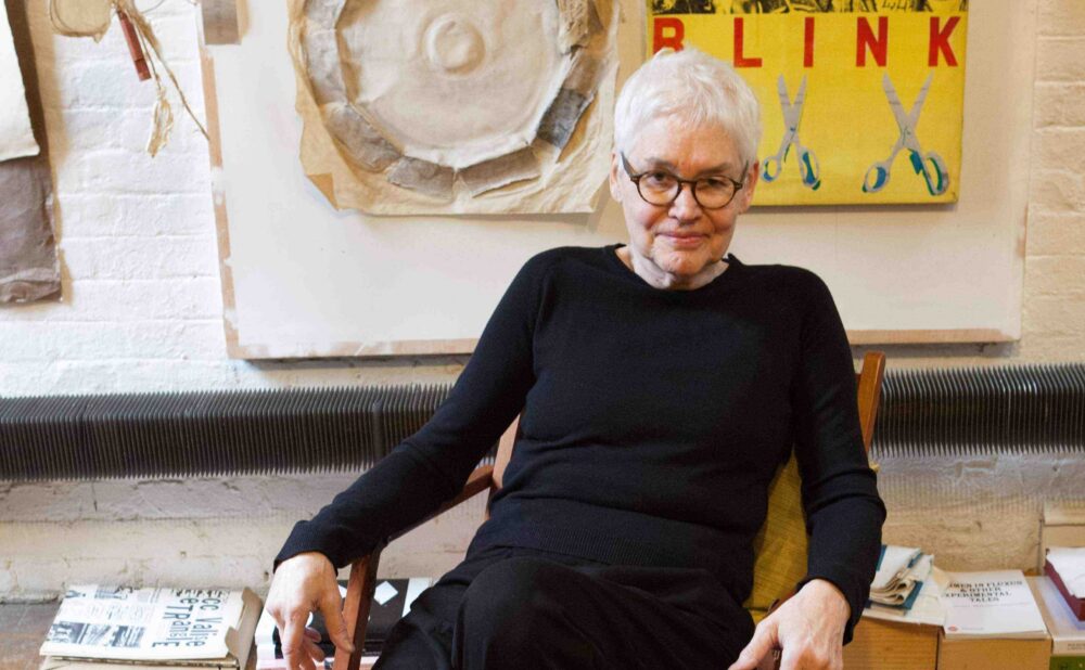 A person wearing black with white hair and glasses sitting in a chair in front of artwork.