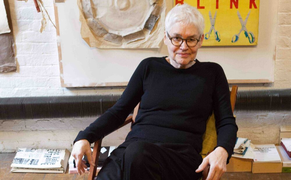 A person wearing black with white hair and glasses sitting in a chair in front of artwork.