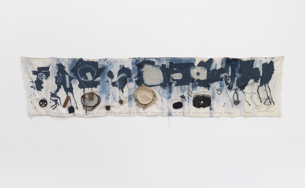 Abstract printed cloth artwork with blue ink patterns and attached objects, hanging on a white background. Alt-text generated by AI.