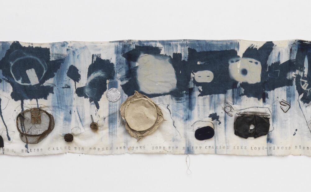 Abstract printed cloth artwork with blue ink patterns and attached objects, hanging on a white background. Alt-text generated by AI.