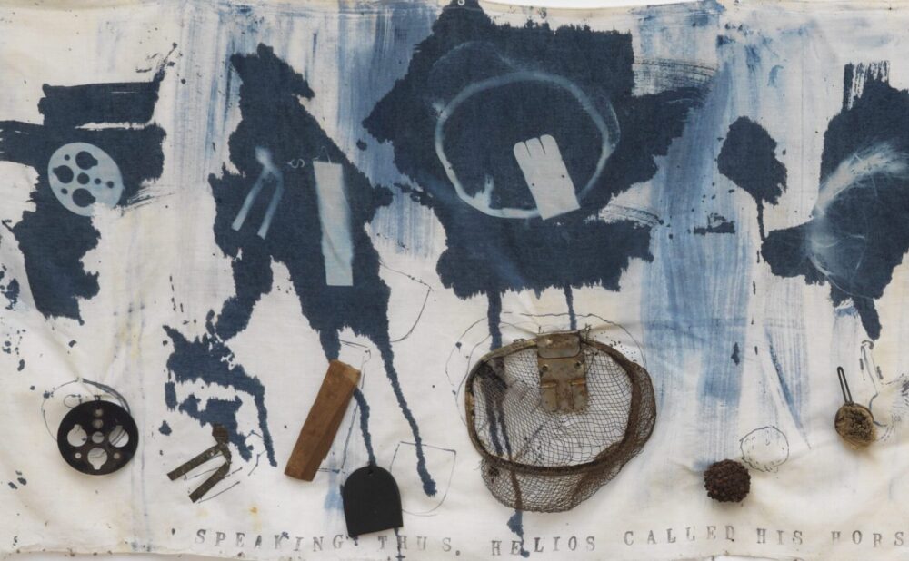 Detail of abstract printed cloth artwork with blue ink patterns and attached objects, hanging on a white background. Alt-text generated by AI.