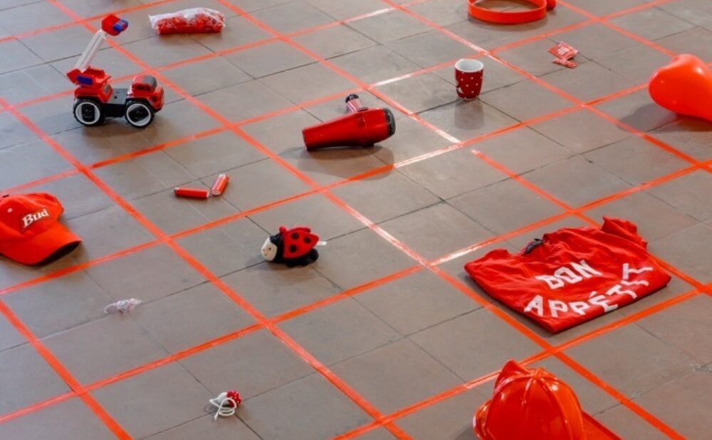 A tiled floor featuring a red grid with an arrangement of various red objects, including a toy car, hats, and containers.