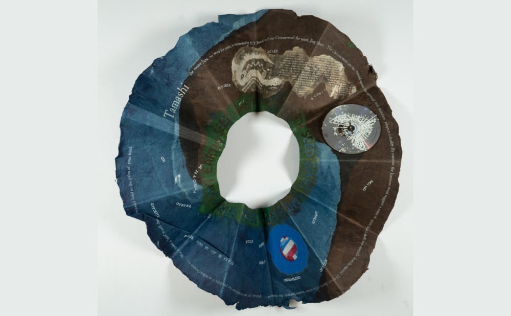 A circular, textured paper sculptural object, with gradients of blue and brown, featuring scattered text. Alt-text generated by AI.