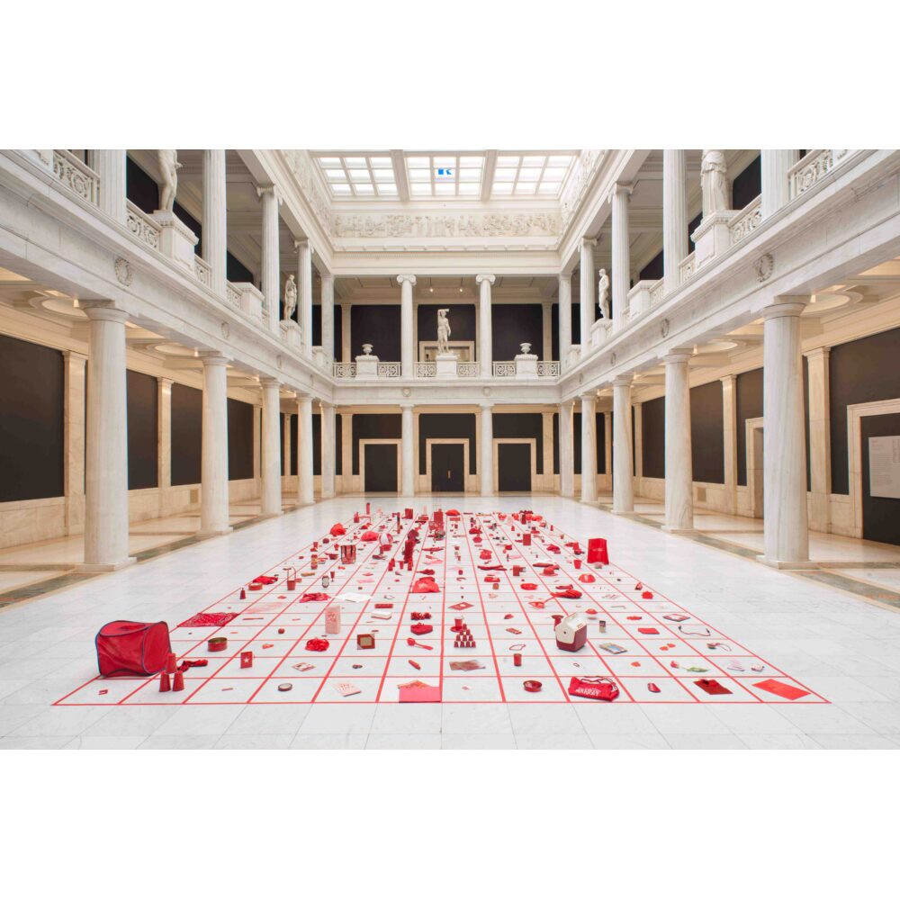 An art installation of red objects arranged in a grid in a classical marble gallery. Alt-text generated by AI.