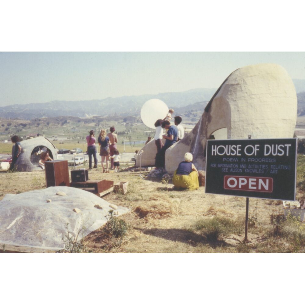 Outdoor art installation with people interacting near dome-like structures and a sign reading "House of Dust" against a hilly landscape. Alt-text generated by AI.  Transcribed Text:  HOUSE OF DUST POEM IN PROGRESS FOR INFORMATION AND ACTIVITIES RELATING SEE ALISON KNOWLES / ART OPEN