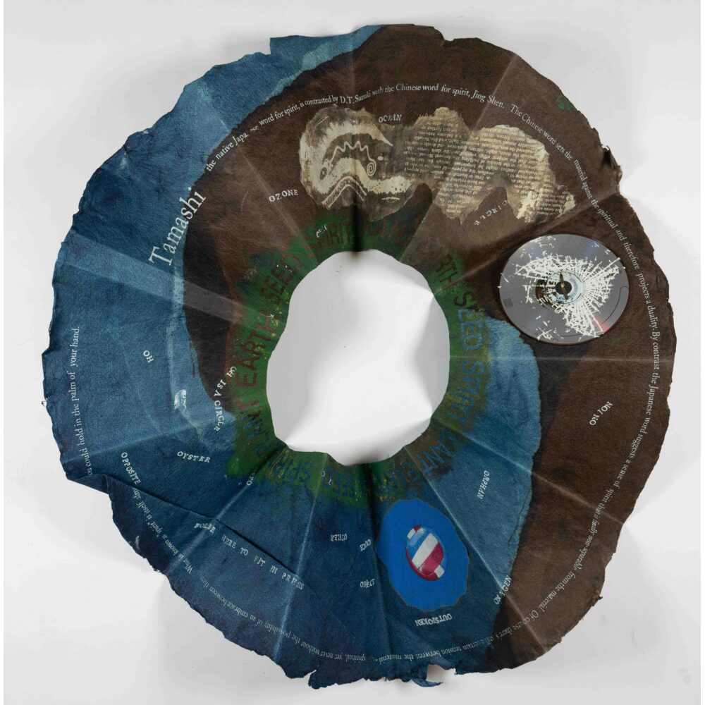 A circular, textured paper sculptural object, with gradients of blue and brown, featuring scattered text. Alt-text generated by AI.