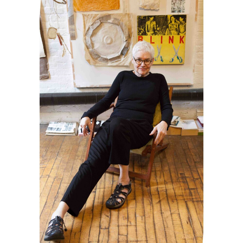 A person with white hair in black clothing seated on a wooden chair in an art studio. Alt-text generated by AI.