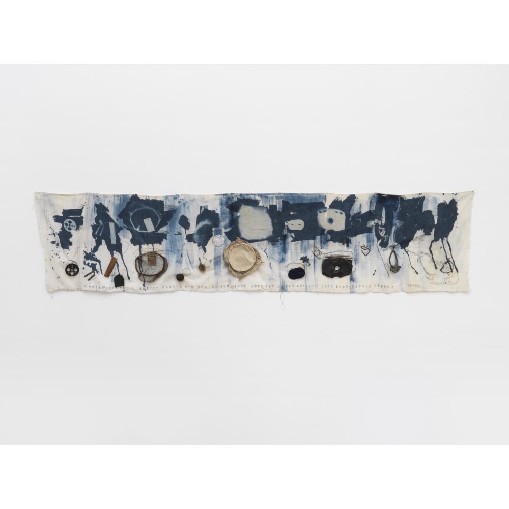 Abstract printed cloth artwork with blue ink patterns and attached objects, hanging on a white background. Alt-text generated by AI.