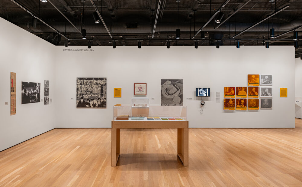 Wide view of a gallery showing photographs, posters, archival materials, and canvases arranged on white walls around a central glass display case. Alt text generated by AI.