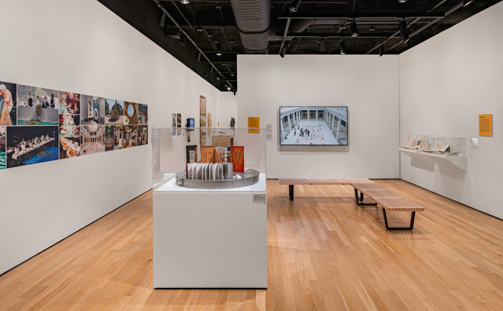 Contemporary museum gallery displaying photographs, artifacts in glass cases, a wall-mounted video screen, and a wooden bench. Alt text generated by AI.