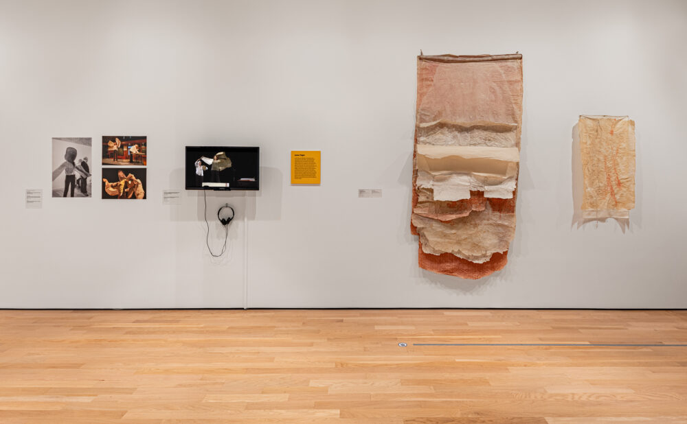 Museum gallery installation with photographs, a video monitor and headphones, wall text, and two large layered paper artworks. Alt text generated by AI.