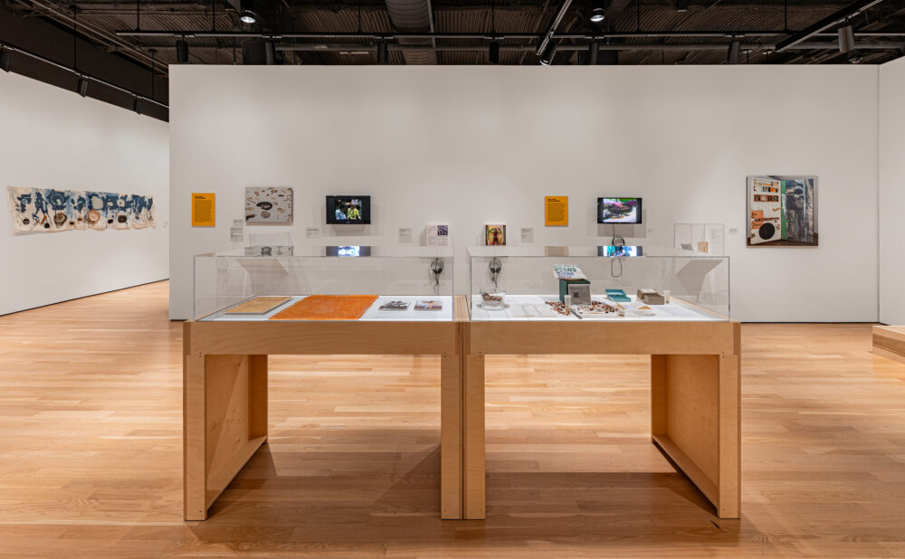 Wide view of a contemporary museum gallery featuring two glass display cases, wall-mounted artworks, video monitors, and interpretive materials. Alt text generated by AI.