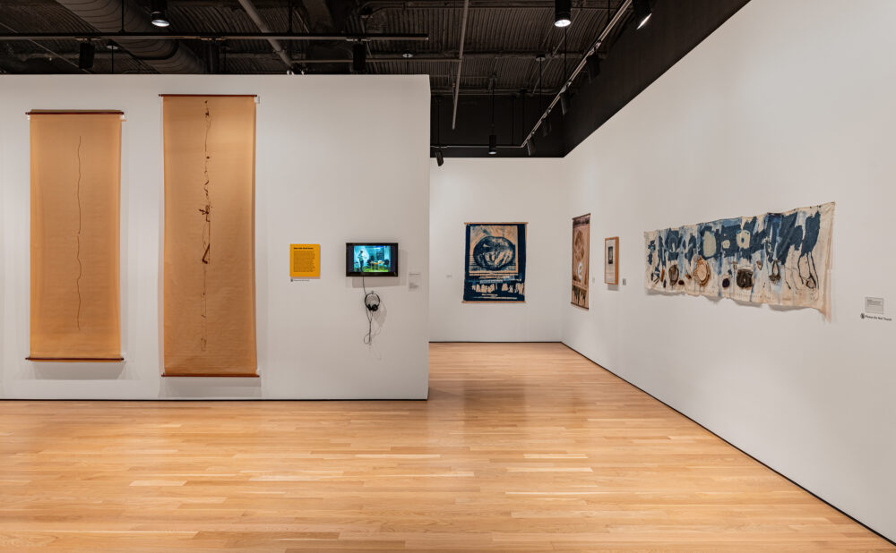 Interior of a contemporary art gallery displaying hanging paper works, framed art, a video monitor, and a large blue-and-beige abstract textile.