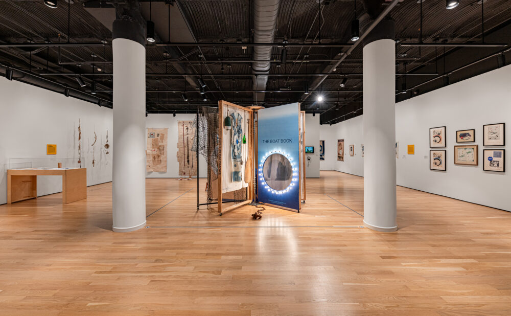 Wide view of a contemporary art gallery with a central boat-themed installation, framed artwork, display cases, white columns, and an exposed industrial ceiling. Alt text generated by AI.
