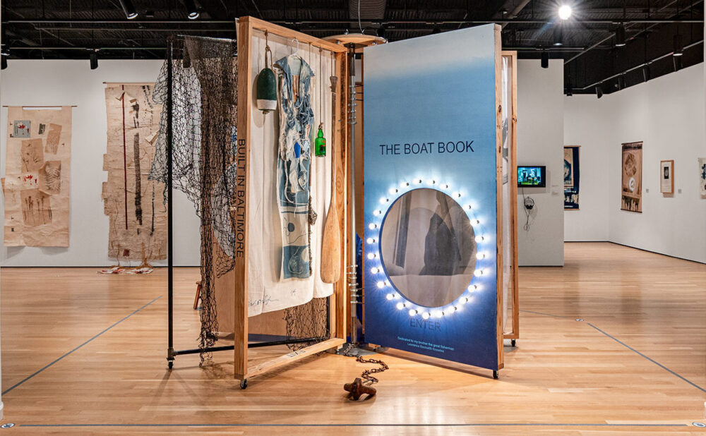 Wide view of a contemporary art gallery with a central boat-themed installation, framed artwork, display cases, white columns, and an exposed industrial ceiling. Alt text generated by AI.