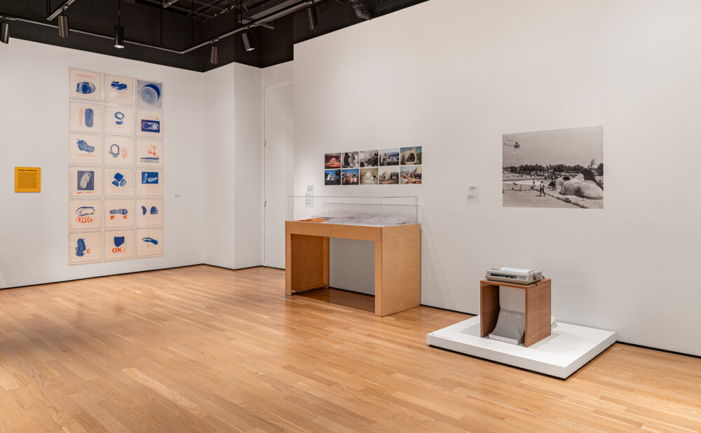 Interior of a contemporary gallery displaying framed prints, photographs, documents, and vintage equipment in wooden and glass cases. Alt text generated by AI.