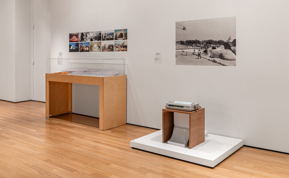 Interior of a contemporary gallery displaying photographs reproductions, a display case with documents, and a dot matrix printer with a scroll of paper.