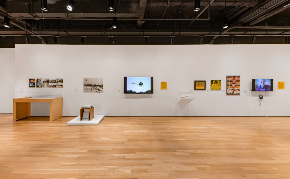 Wide view of a contemporary gallery exhibition with photographs, framed artworks, display cases, and video screens along a white wall.
