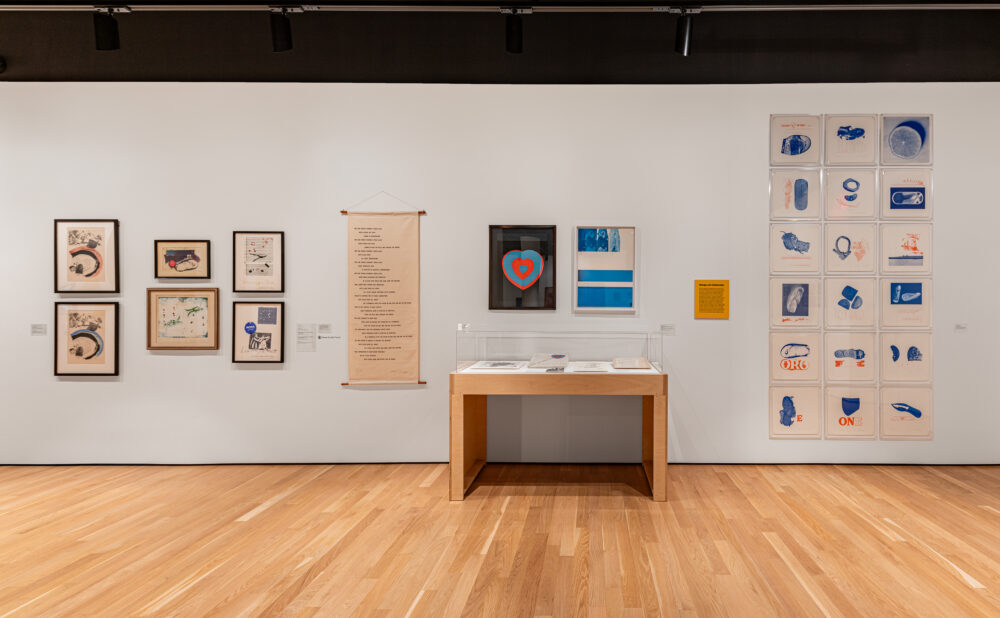 Wide view of a gallery wall displaying framed artworks, a text-covered hanging scroll, a glass display case, and a grid of blue-toned prints.