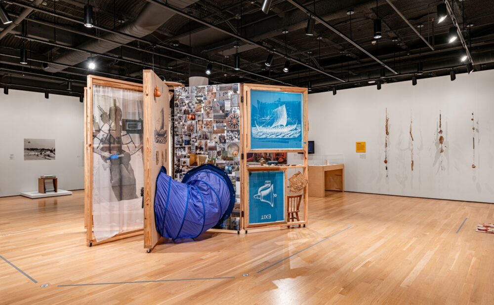 Contemporary gallery featuring a large wheeled wooden art installation covered with photographs, drawings, objects, and blue framed artworks.