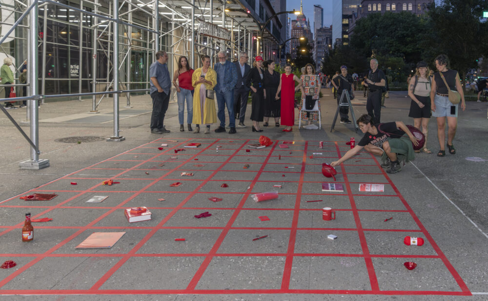 People gather beside a red-taped grid installation scattered with objects on a downtown sidewalk at dusk.