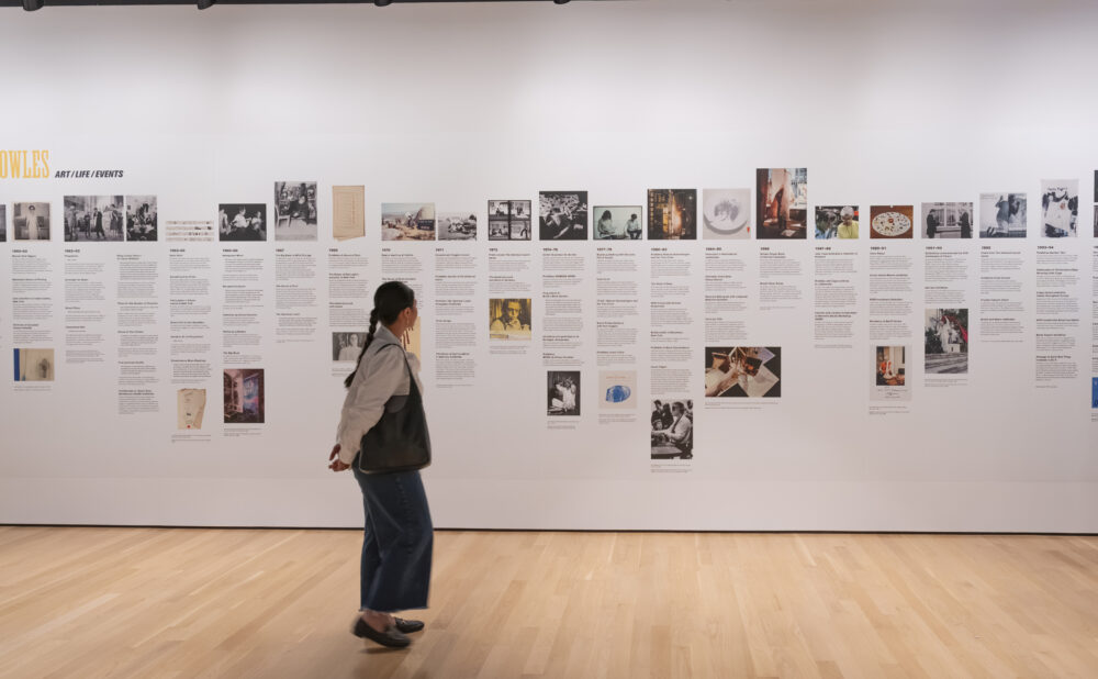 Person walking through a gallery in front of a large chronological exhibition timeline spanning the 1960s through the 1990s. Alt text generated by AI.