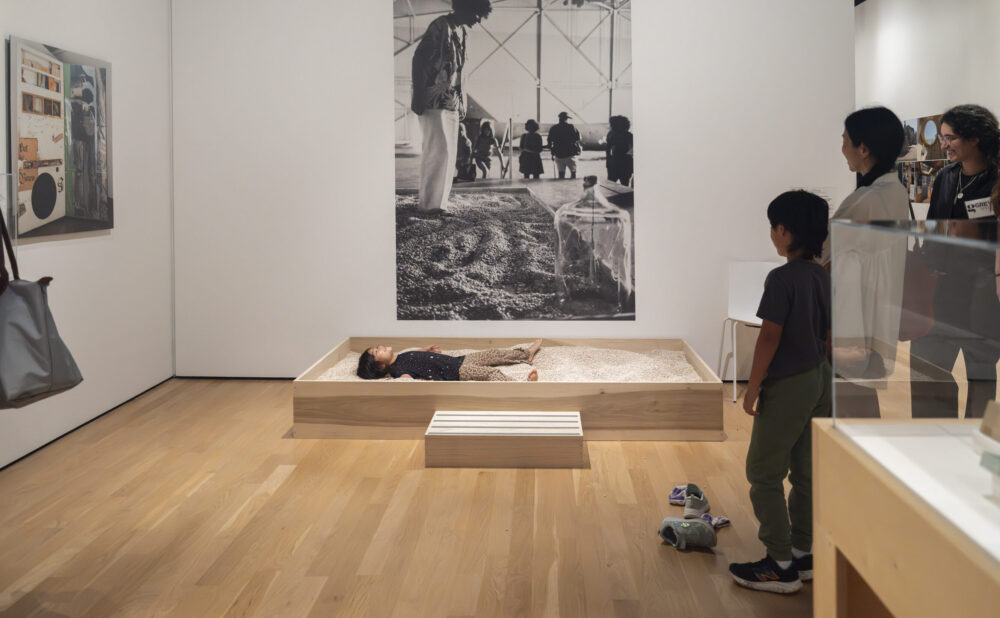 Museum gallery installation with a person lying in a wooden bed of white beans beneath a large black-and-white photograph, while visitors observe.