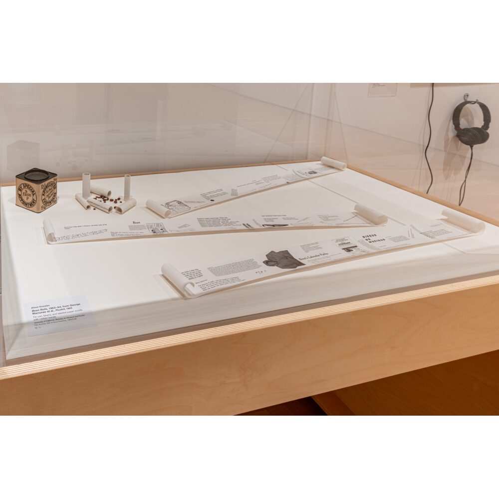 Museum display of Alison Knowles’s Bean Rolls, featuring paper scrolls, beans, a labeled metal can, and headphones on the wall. Alt text generated by AI.