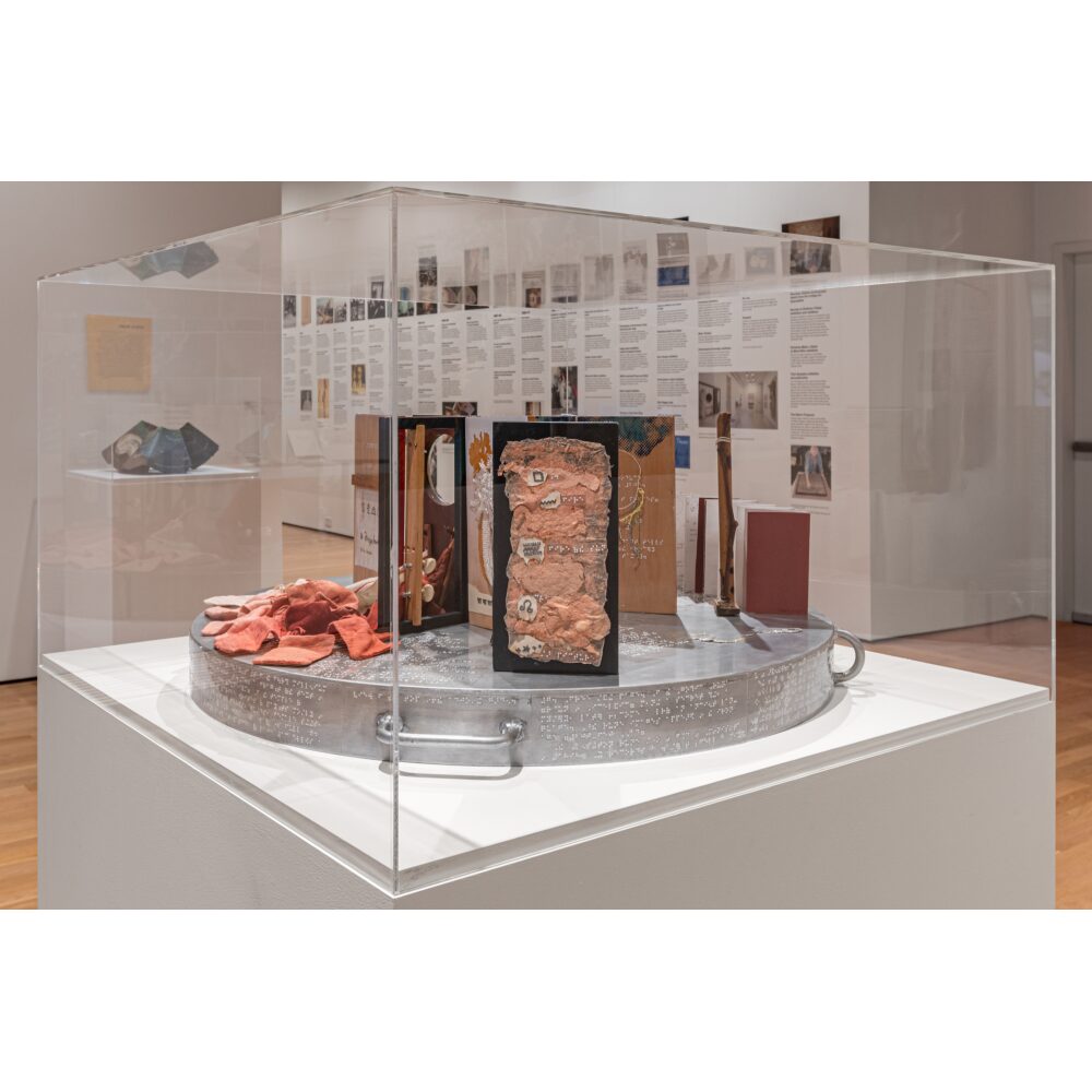 Museum display case containing a tactile mixed-media artwork with fabric, wood, textured panels, and raised-dot markings. Alt text generated by AI.