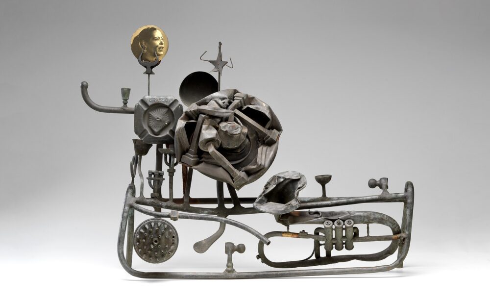 Sculptural assembly of metallic objects including a rose, pipes, and a portrait disk on a gray background.
