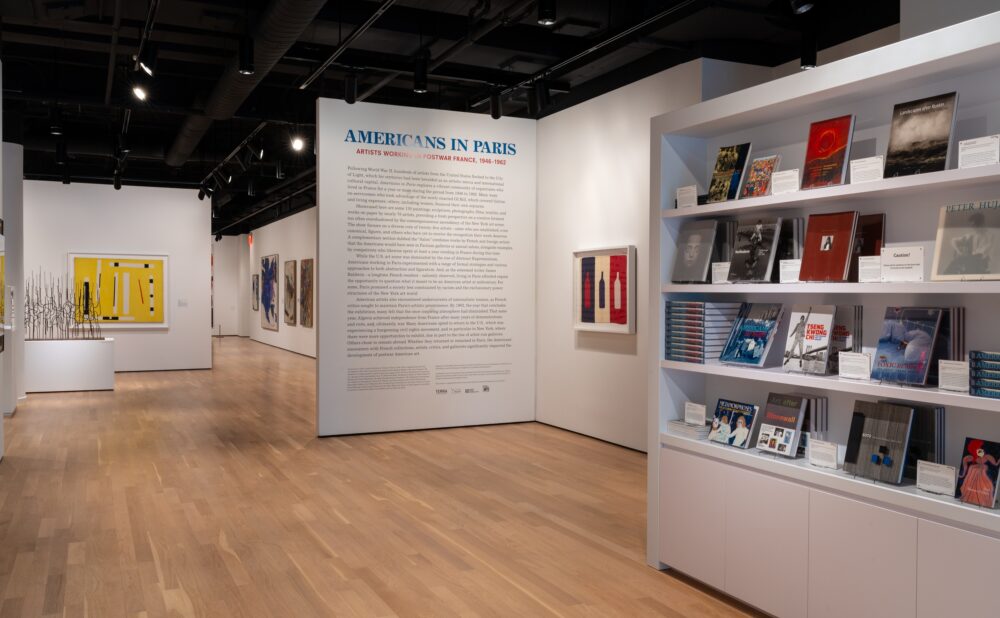 Art gallery exhibition with artworks, a text panel titled "AMERICANS IN PARIS," and shelves of art books.