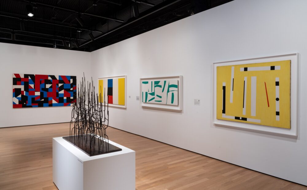 Gallery with abstract paintings and a black, metal sculpture on a pedestal.