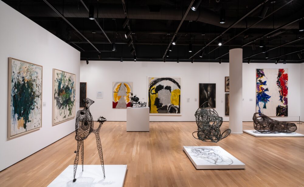 An art gallery featuring abstract paintings and sculptures on a wooden floor with bright lighting.